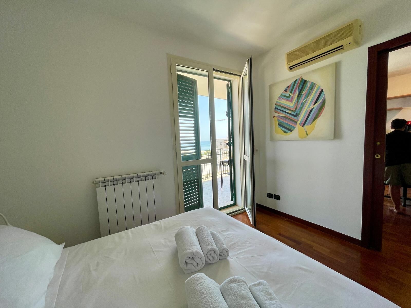 A hotel room with a bed, a radiator, a window with blinds, a door, and a wall with a colorful abstract painting.