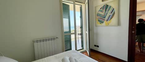 A hotel room with a bed, a radiator, a window with blinds, a door, and a wall with a colorful abstract painting.