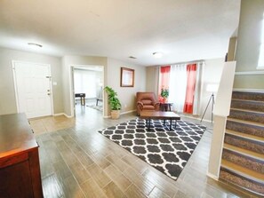 A spacious living room with hardwood flooring, a black and white rug, a brown leather chair, a small table, and a staircase leading to another level. - Fully Loaded 4BR Cul-de-Sac Home with 3 Living Rooms, Game Room, & big backyard (Fort Worth)