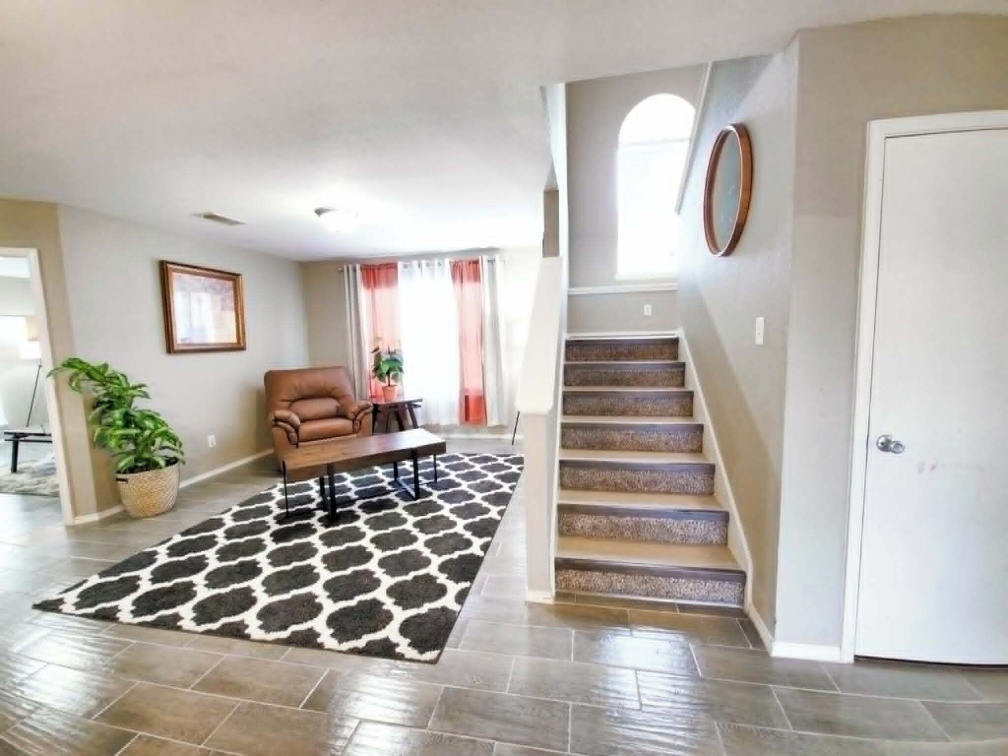 A spacious living room with a brown leather sofa, a coffee table, and a patterned rug. A staircase with a wooden finish leads to an upper level.