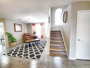 A spacious living room with a brown leather sofa, a coffee table, and a patterned rug. A staircase with a wooden finish leads to an upper level. - Fully Loaded 4BR Cul-de-Sac Home with 3 Living Rooms, Game Room, & big backyard (Fort Worth)
