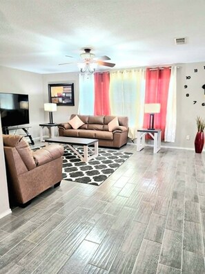 A modern living room with a sofa, armchair, and coffee table, a flat-screen TV, and a ceiling fan. - Fully Loaded 4BR Cul-de-Sac Home with 3 Living Rooms, Game Room, & big backyard (Fort Worth)