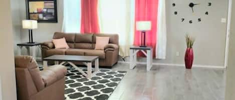 Un salon avec un canapé, une table basse et un tapis. On y trouve un ventilateur de plafond, une horloge au mur et un vase rouge posé sur le sol.