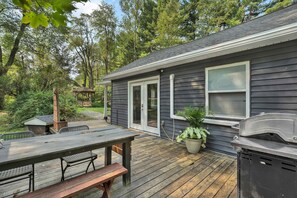 Eine Terrasse mit Grill und Tisch, umgeben von einem Garten und einem Haus mit dunkel verputzter Fassade.
