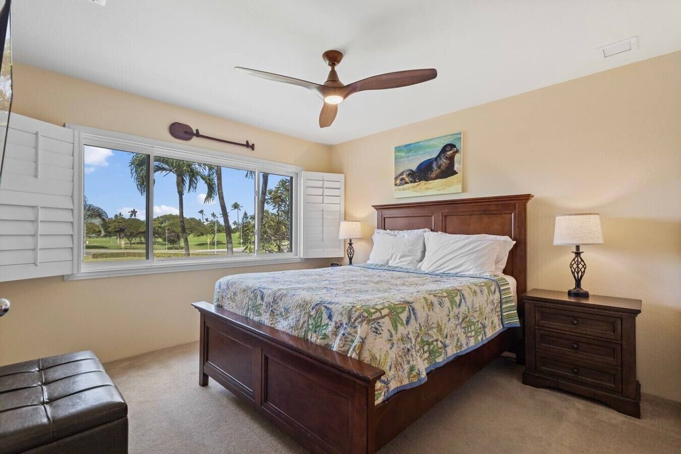 A bedroom with a bed, a ceiling fan, a window with shutters, a nightstand with a lamp, and a painting on the wall.