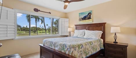 A bedroom with a bed, a ceiling fan, a window with shutters, a nightstand with a lamp, and a painting on the wall.