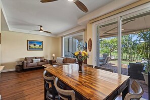 A spacious living area with a wooden dining table, a sofa set, and a large window offering a view of the outdoors. - Wake Up to Golf Course Views & Resort Amenities (Kapolei)