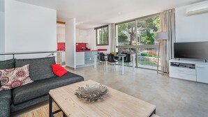 Una sala de estar moderna con sofá, una mesa de centro y televisor. Cuenta con una zona de cocina con encimera roja y una mesa de comedor con sillas. La habitación tiene ventanas grandes con cortinas.