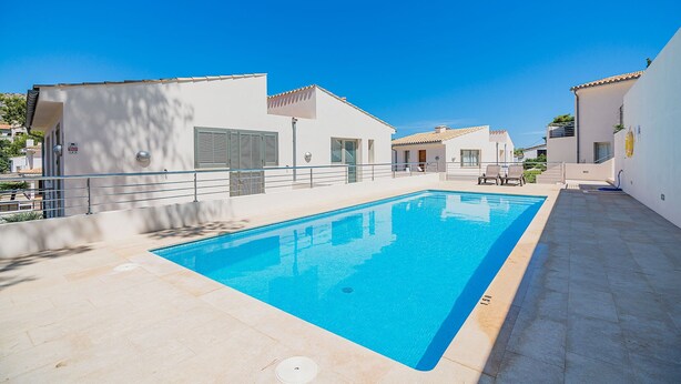 Una piscina rectangular con solárium empedrado, rodeada de edificios blancos con balcones y un cielo azul despejado.