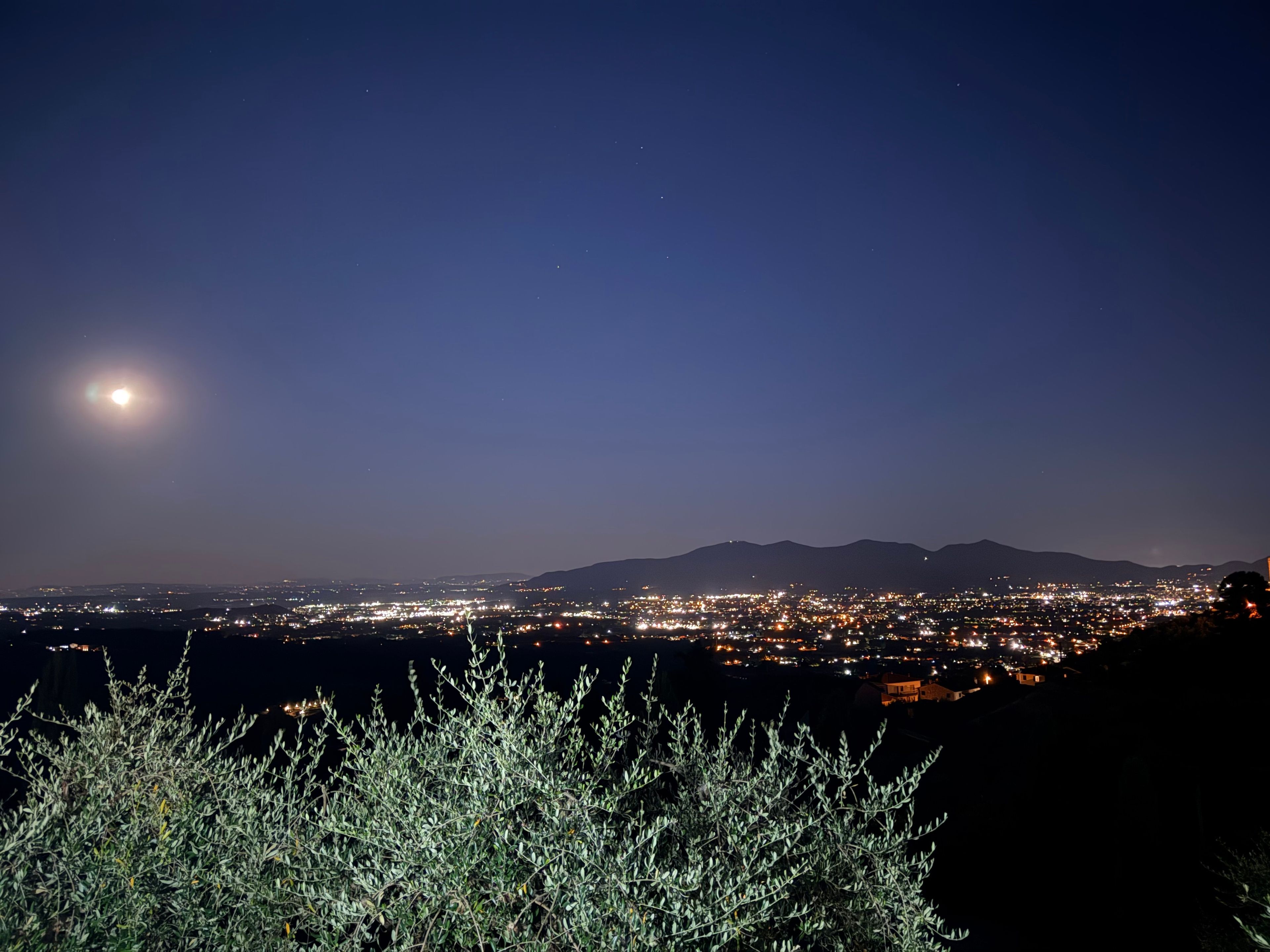 A night view of a city with a mountain range in the background and a crescent moon.