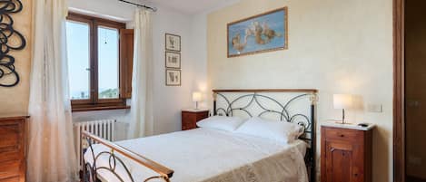 A bedroom with a bed, two bedside tables, a window with curtains, a radiator, and a painting on the wall.