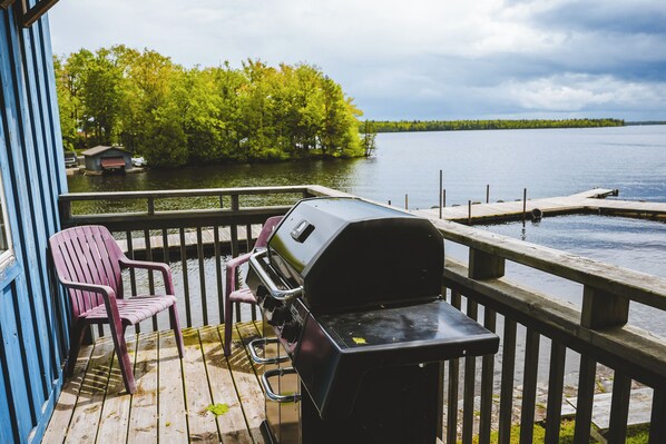 A wooden deck with a grill and two chairs, overlooking a calm lake with trees and a dock in the background.