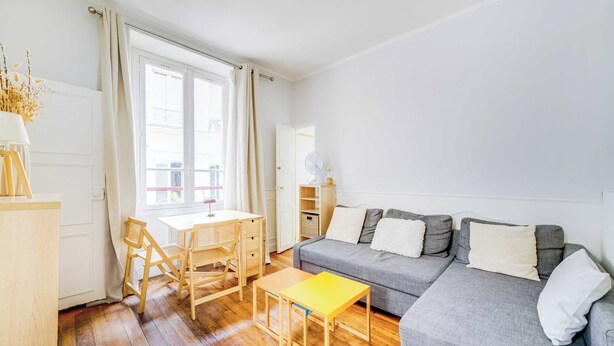 Un salon lumineux avec un plancher en bois, un canapé gris, une petite table jaune et un bureau en bois avec des chaises.