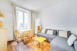 Een lichte woonkamer met houten vloer, een grijze bank, een klein geel tafeltje en een houten bureau met stoelen.