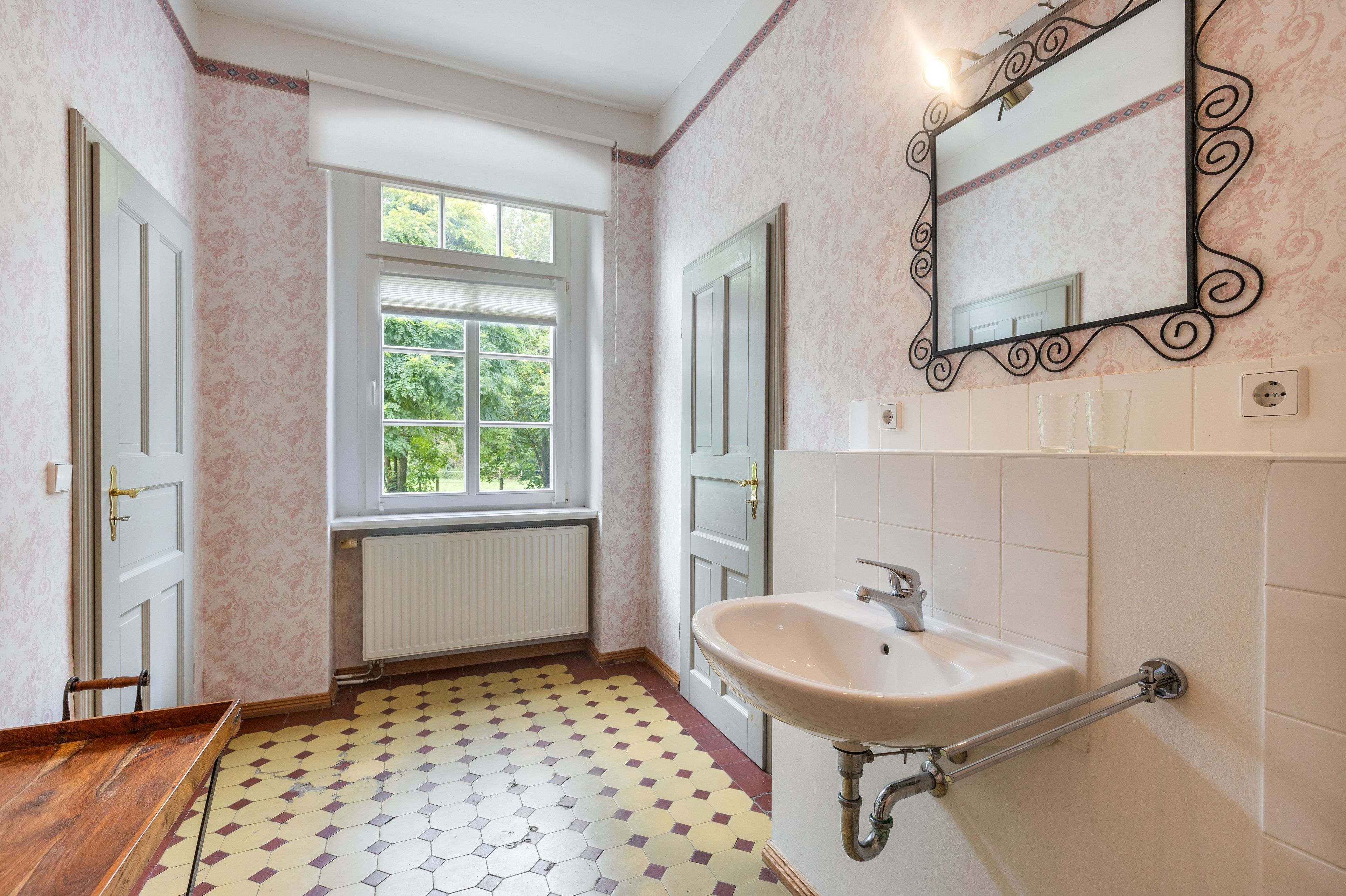 A bathroom with a white sink, a mirror, a window, and patterned wallpaper.