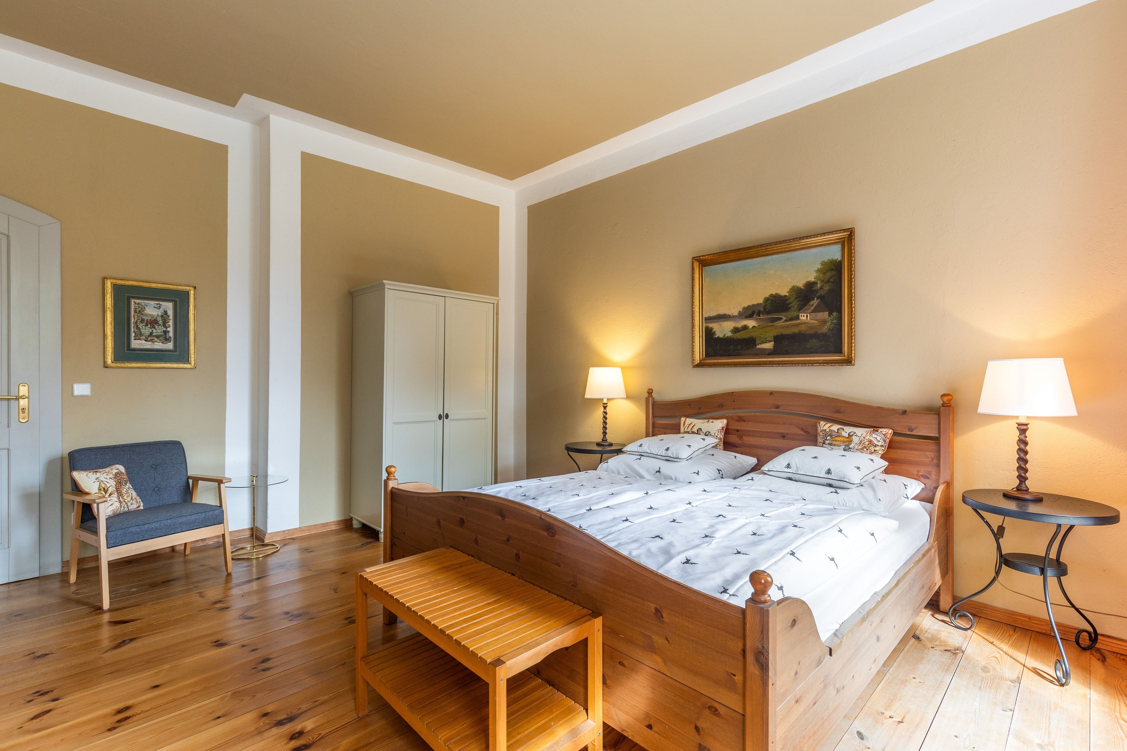 Un dormitorio con una cama de madera, un banco, una silla, un armario, un cuadro y dos lámparas.