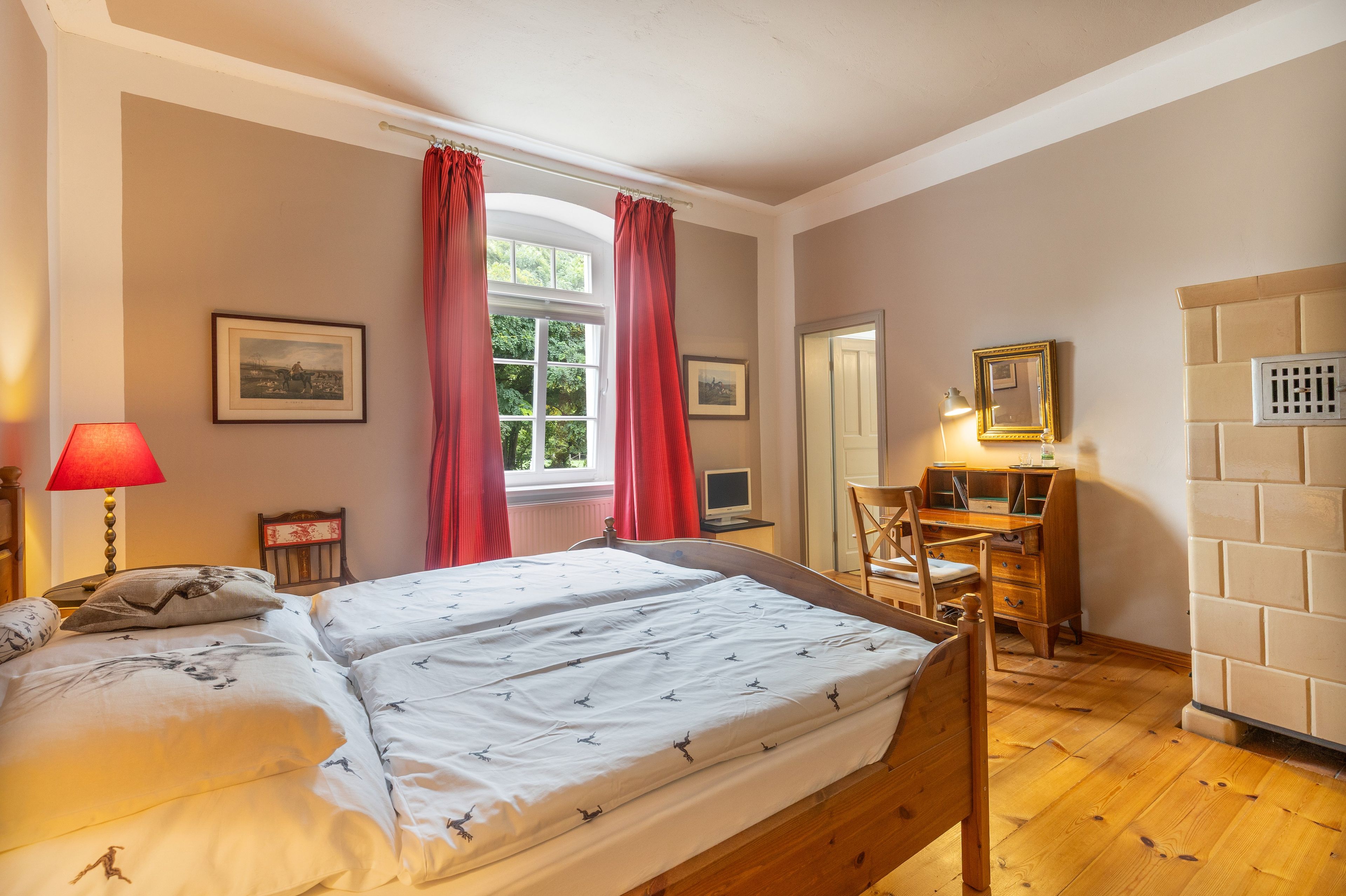 Ein Schlafzimmer mit Doppelbett, Schreibtisch mit Stuhl, Fenster mit Vorhängen und Heizkörper.