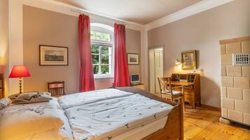 Ein Schlafzimmer mit Doppelbett, Schreibtisch mit Stuhl, Fenster mit Vorhängen und Heizkörper.