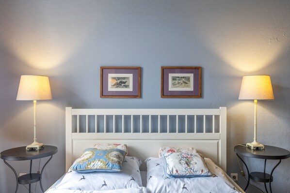 A bedroom with a white headboard, two bedside tables, two lamps, and two framed pictures on the wall.
