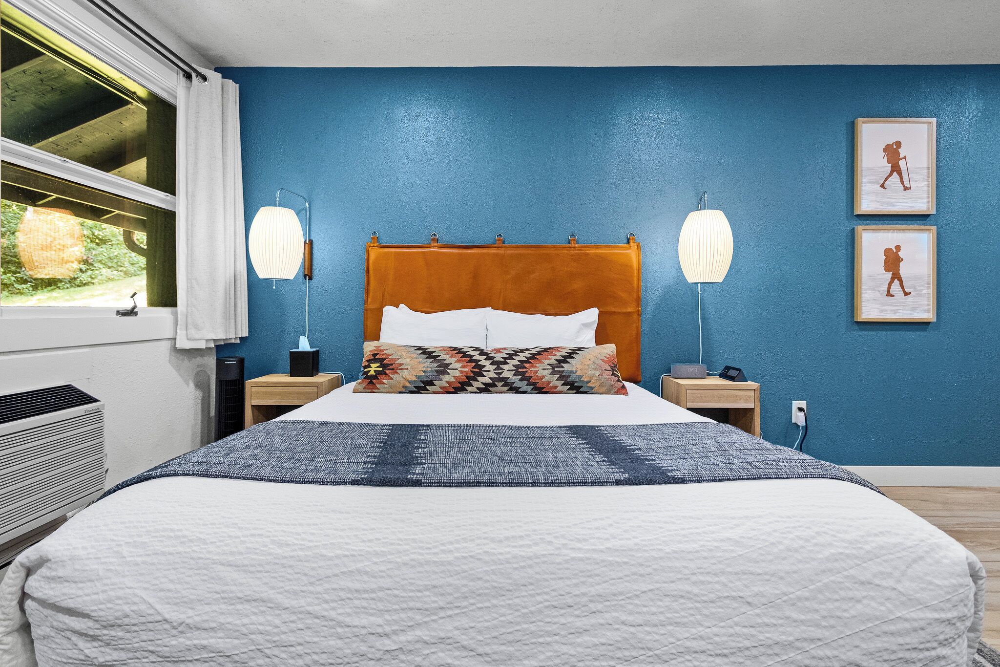 A bedroom with a bed, bedside lamps, a nightstand, a window with a view, and a wall with framed pictures.