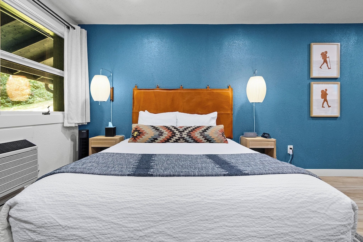 A bedroom with a bed, bedside lamps, a nightstand, a window with a view, and a wall with framed pictures.