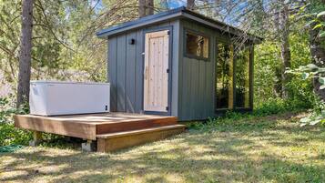 Un piccolo capanno di legno con una porta e una finestra, un elettrodomestico bianco su una piattaforma in legno, e una zona erbosa con alberi.