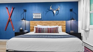 Un dormitorio con cama, cabecera de madera, pared azul, cráneo de ciervo montado y decoración roja en forma de X.