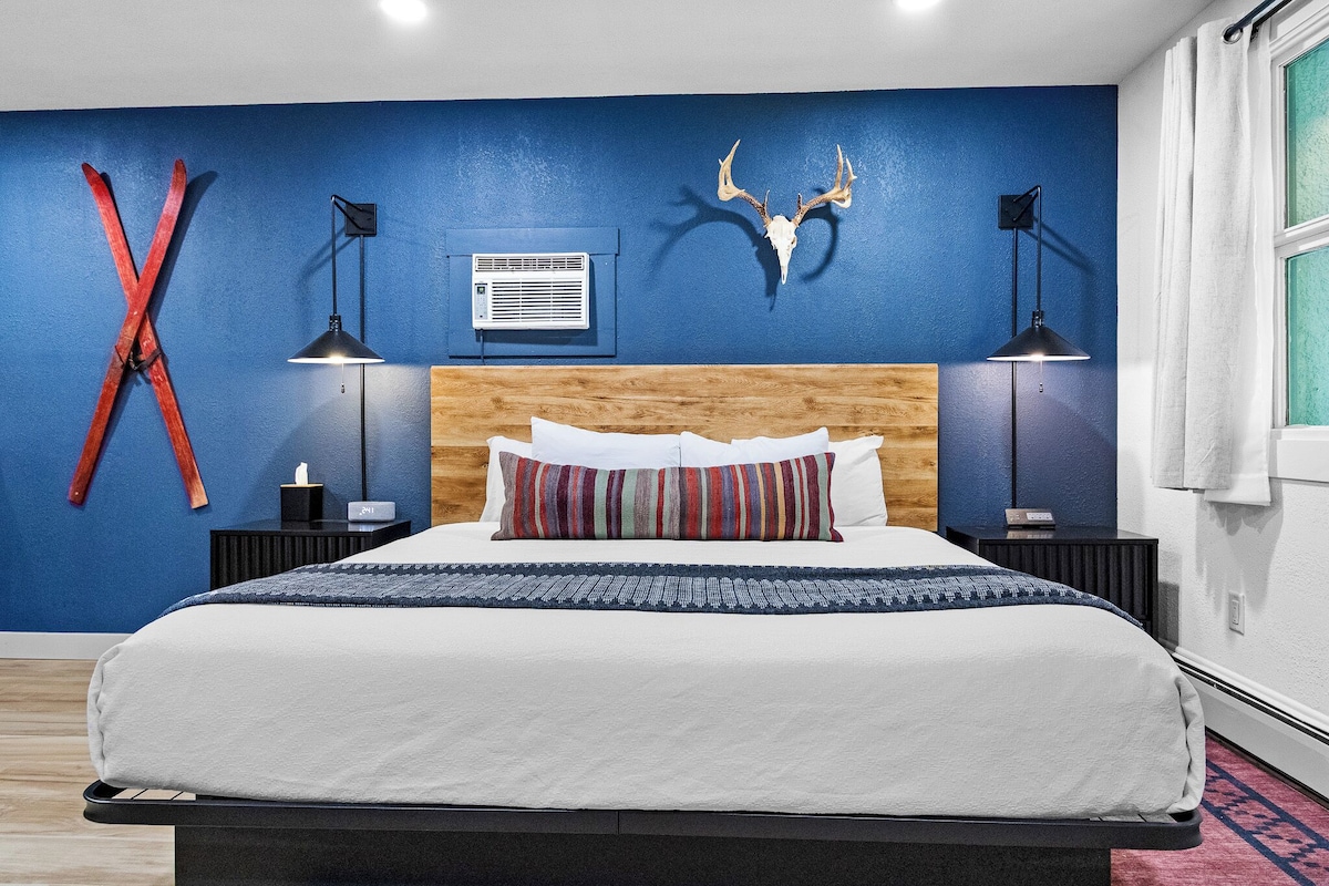 A bedroom with a bed, a wooden headboard, a blue wall, a mounted deer skull, and a red X-shaped decoration.