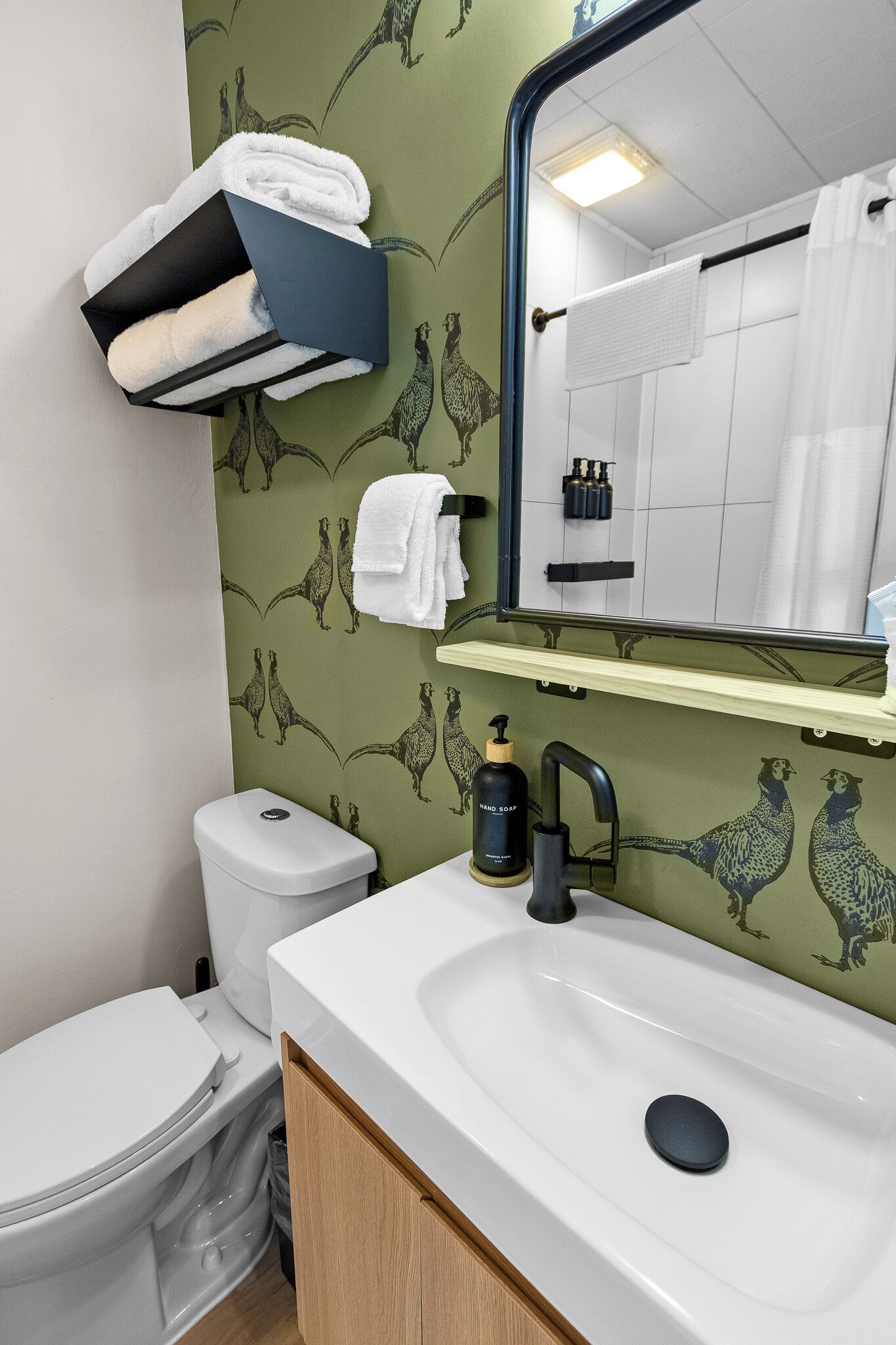 Une salle de bain avec un lavabo blanc, un miroir et des toilettes. Des serviettes sont suspendues sur un porte-serviettes et une étagère se trouve au-dessus du lavabo.
