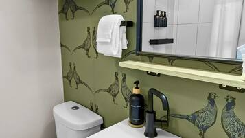 Une salle de bain avec un lavabo blanc, un miroir et des toilettes. Des serviettes sont suspendues sur un porte-serviettes et une étagère se trouve au-dessus du lavabo.