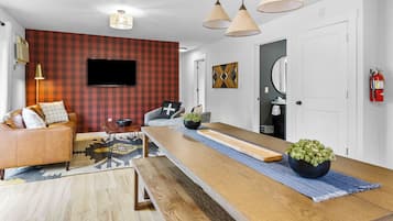 Una sala de estar moderna con un sofá, una mesa de centro, un televisor montado en la pared y una zona de comedor con una mesa de madera larga.