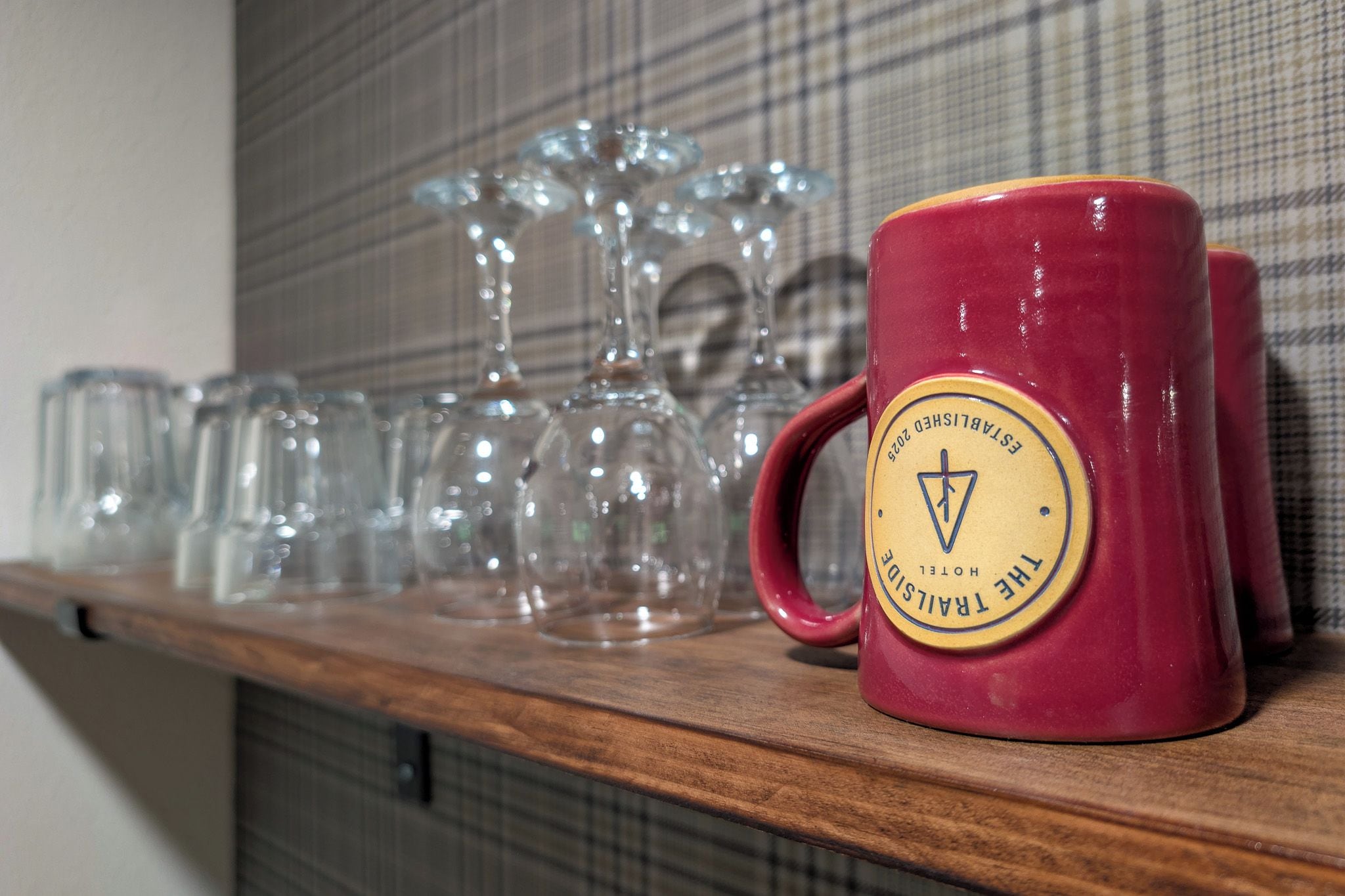 Una taza roja con el logotipo de un hotel sobre una estantería junto a vasos de vidrio.