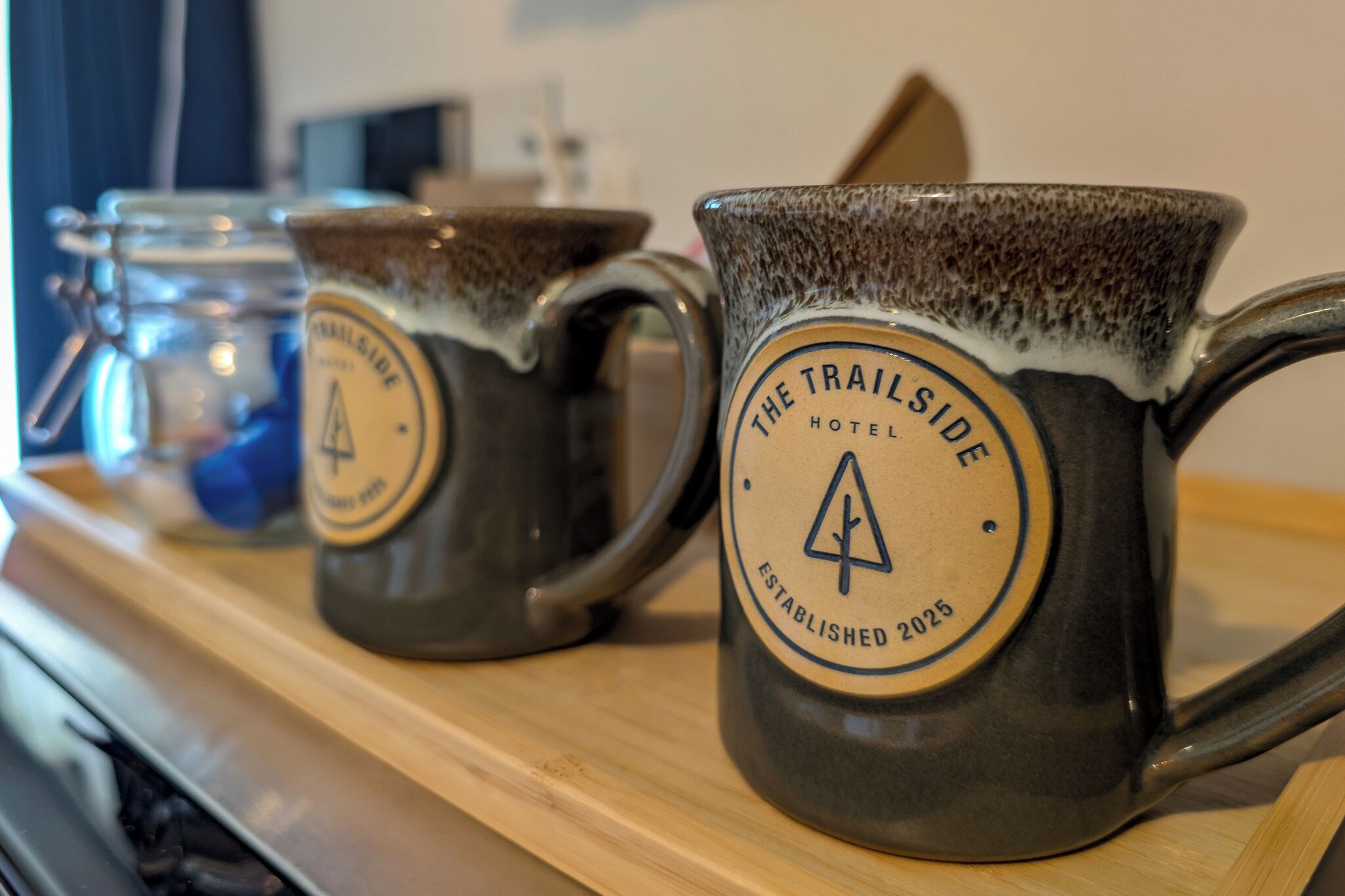 Two mugs with 'The Trailside Hotel' logo and a mountain symbol, placed on a wooden surface.