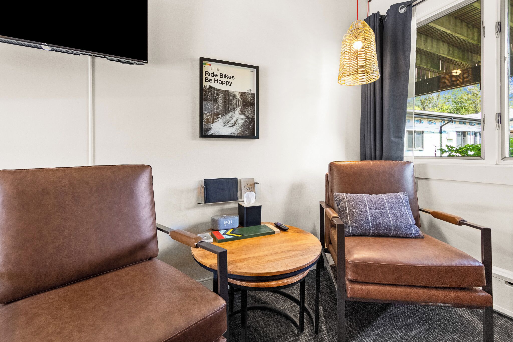 A room with two brown leather chairs, a small round table with a book and a remote control, a wall-mounted TV, a framed picture with text, and a window with curtains.