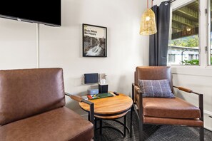 A room with two brown leather chairs, a small round table with a book and a remote control, a wall-mounted TV, a framed picture with text, and a window with curtains.