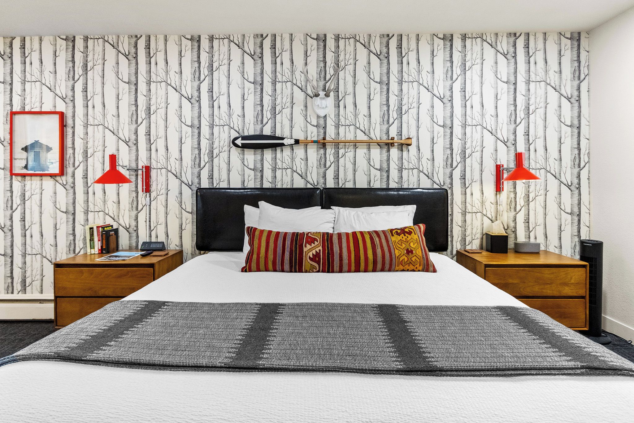 A bedroom with a bed, two nightstands, a red lamp, a wall clock, a framed picture, and a wall-mounted shelf.
