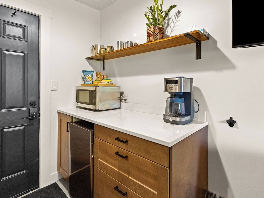 A compact kitchen with a microwave, coffee maker, and a small shelf with a plant and snacks.