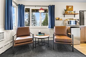 A room with two leather chairs, a small table, a window with curtains, a hanging light, and a kitchen area with appliances.