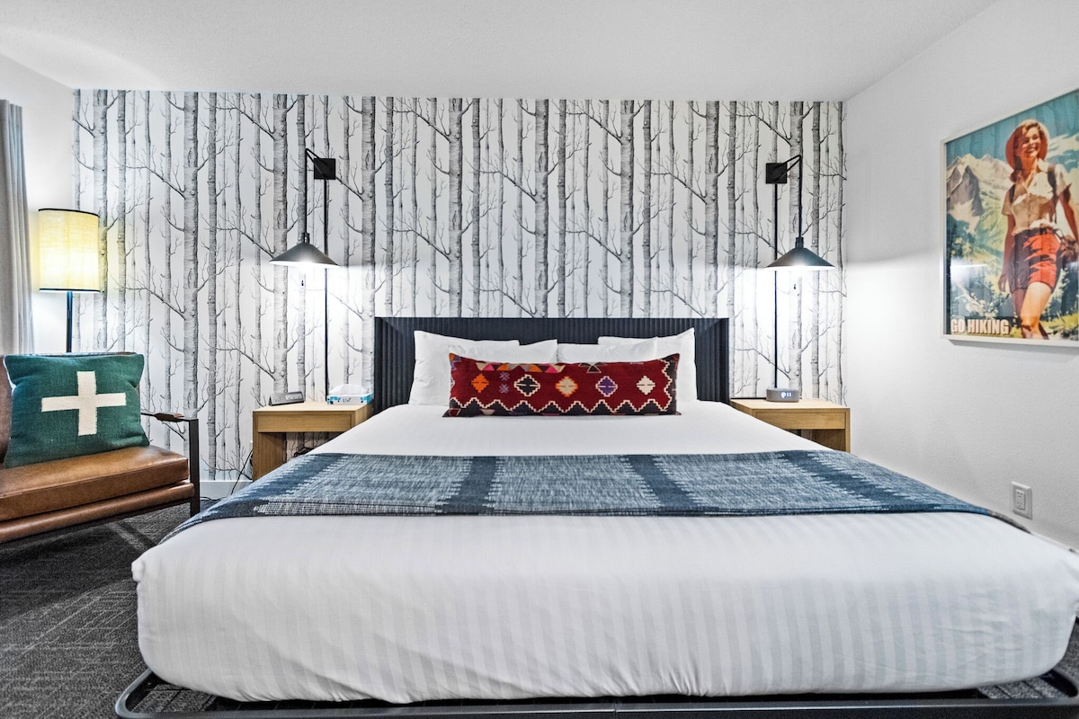 A hotel room with a large bed, two bedside tables, a nightstand, a wall-mounted lamp, a painting, and a window with curtains.