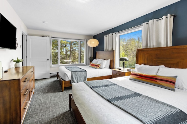 Habitación de hotel con dos camas, un escritorio, un televisor y ventanas grandes con cortinas.