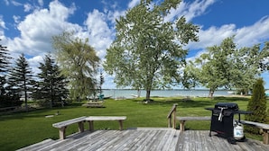 A wooden deck with picnic tables and a grill, overlooking a lake with trees and a clear sky.