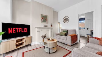 Una sala moderna con un televisor de pantalla plana mostrando 'NETFLIX', una chimenea, un sofá y un comedor a la vista a través de una puerta abierta.