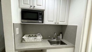 A compact kitchen with white cabinetry, a built-in microwave, a sink, and a countertop with a dish rack. - Shine Suites Balcony Studio (Miami)