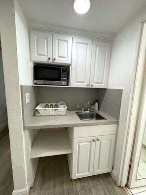 A compact kitchen with white cabinetry, a built-in microwave, a sink, and a countertop with a dish rack. - Shine Suites Balcony Studio (Miami)
