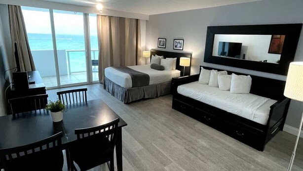 A hotel room with a bed, a desk, a chair, a dining table, and a large window with a view. - Shine Suites Balcony Studio (Miami)