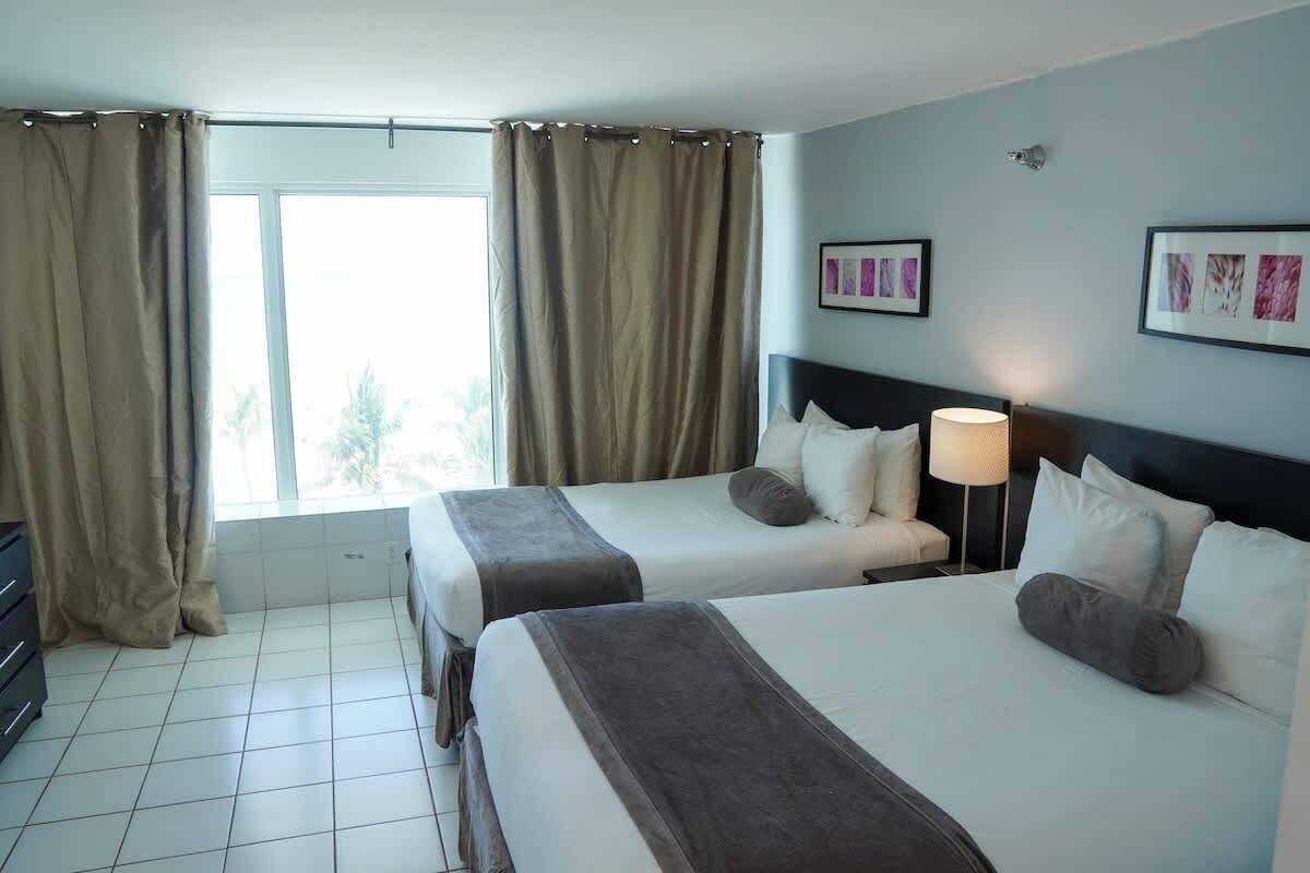 A hotel room with two beds, a window with curtains, a wall-mounted lamp, and framed pictures.
