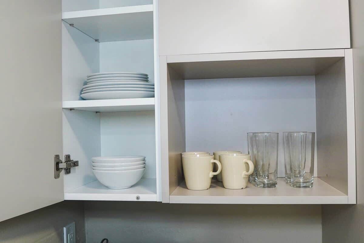 A kitchen cabinet with white shelves, containing stacked white plates and bowls on the upper shelf, and two beige mugs and three clear glasses on the lower shelf.