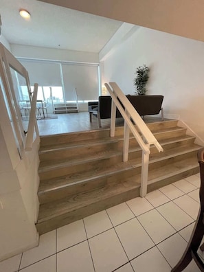 Eine Treppe mit Holzboden und weißen Geländern führt zu einem Raum mit einem schwarzen Stuhl und einer Pflanze.