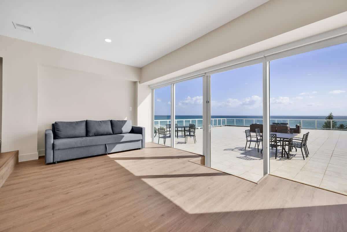 A modern living room with a grey sofa, a wooden floor, and a large sliding glass door leading to a balcony with outdoor furniture.
