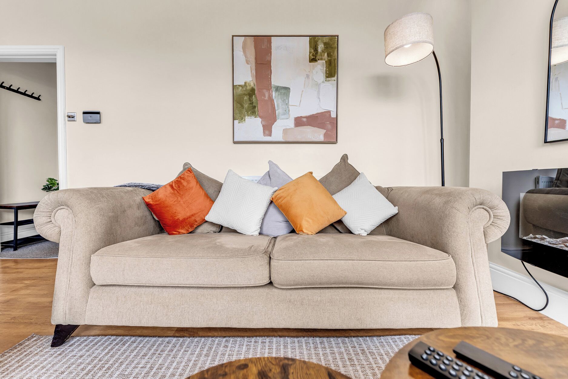 A beige sofa with multiple cushions in various textures and colors, a floor lamp, a painting on the wall, and a remote control on a wooden table.